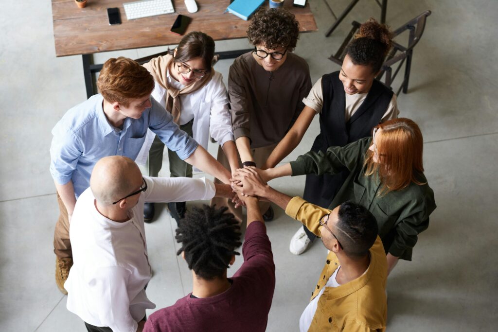 A diverse group of seven people standing in a circle, each placing one hand in the center to form a unified stack, viewed from above.